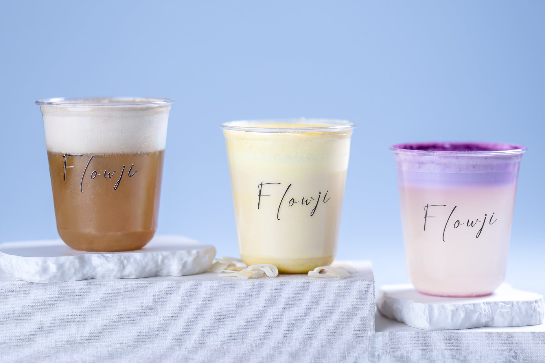 Three Flowji signature cloud drinks displayed in branded plastic cups, featuring Hojicha, Banana Milk, and Coconut Ube flavors on elegant white stands.