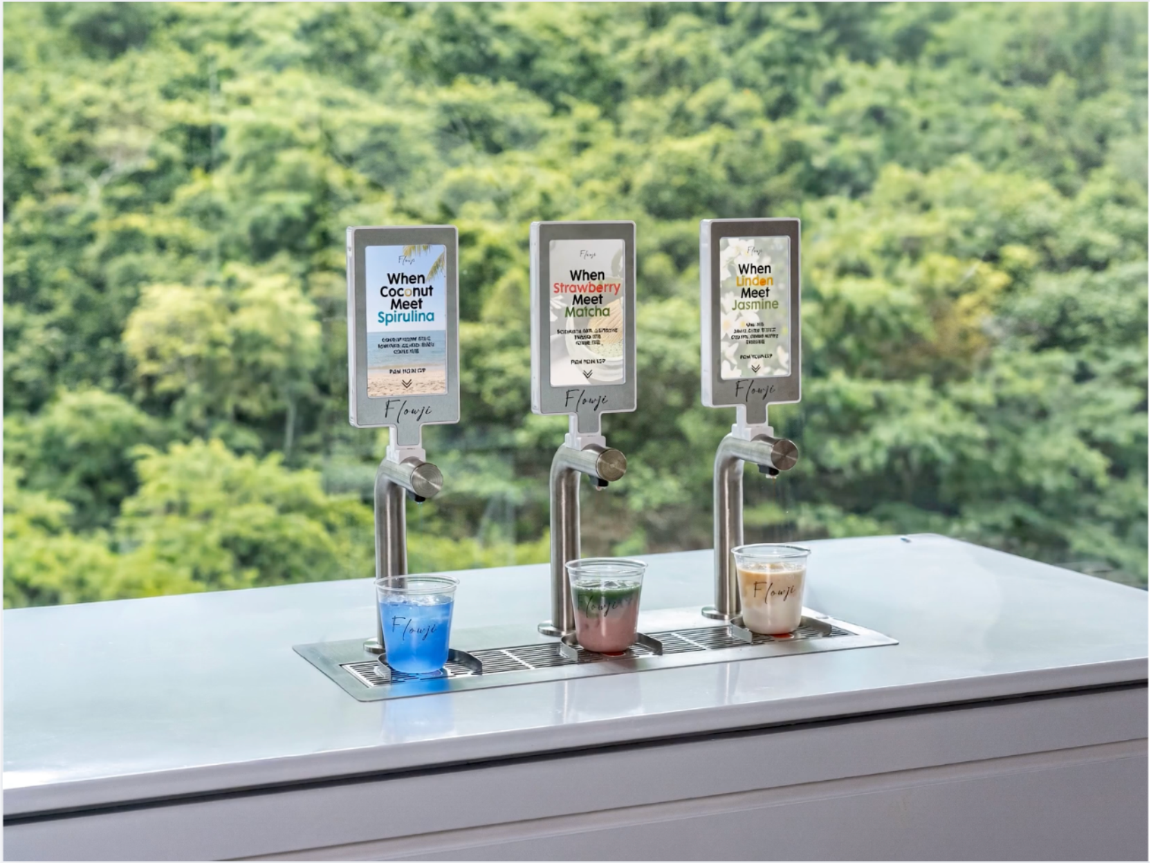 Three Flowji beverage tap dispensers on a stainless steel counter with small cups of blue, pink, and beige drinks in front, set against a blurred green forest background.