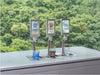 Three Flowji beverage tap dispensers on a stainless steel counter with small cups of blue, pink, and beige drinks in front, set against a blurred green forest background. The right dispenser is pouring.