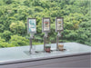 Three Flowji beverage tap dispensers on a stainless steel counter with small cups of blue, pink, and beige drinks in front, set against a blurred green forest background. The left dispenser is pouring.