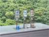 Three Flowji beverage tap dispensers on a stainless steel counter with small cups of blue, pink, and beige drinks in front, set against a blurred green forest background. The middle dispenser is pouring.